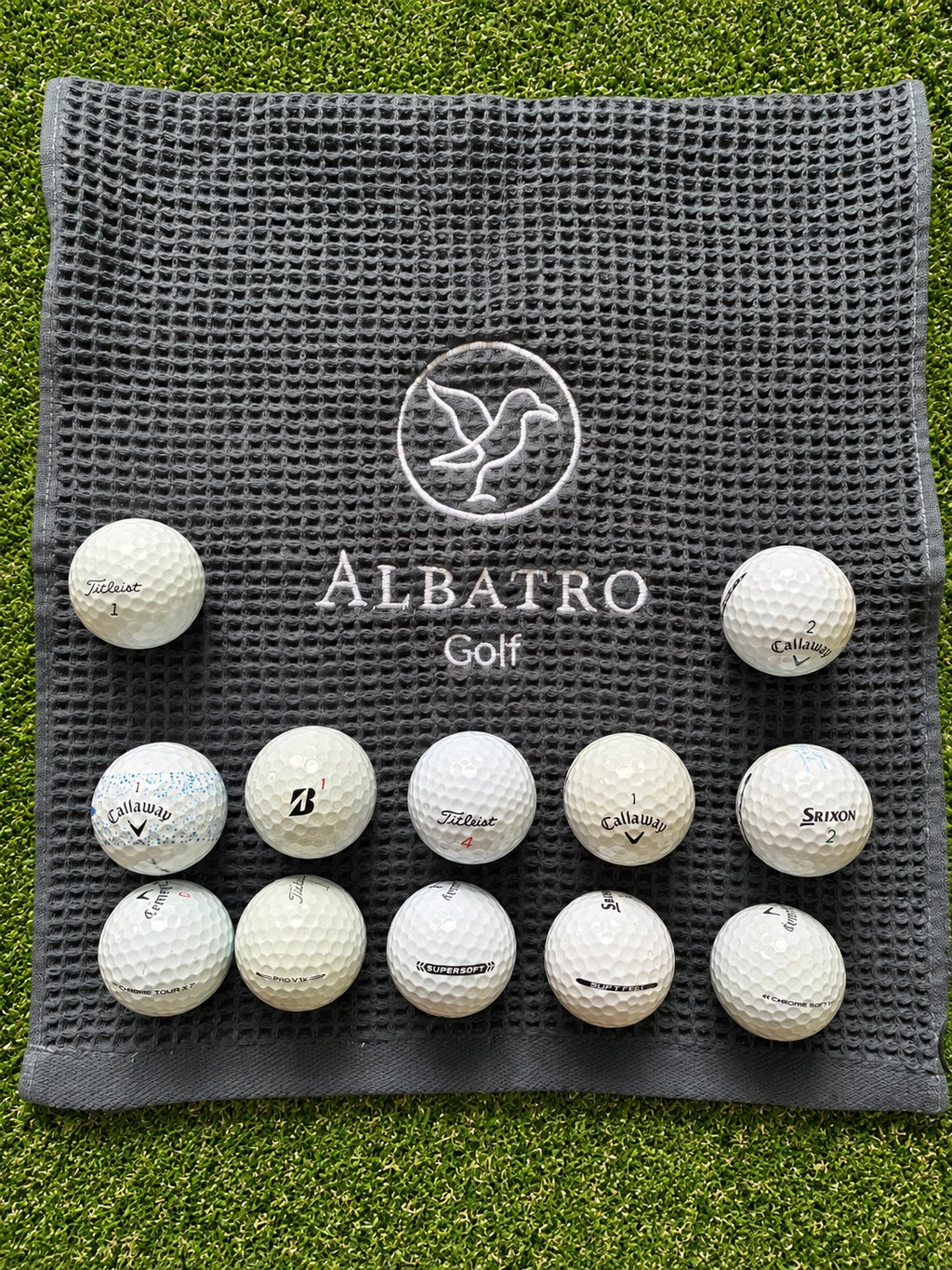 12 pelotas de golf usadas grado AAA Titleist, Callaway, Bridgestone y Srixon sobre toalla Albatro Golf en pasto